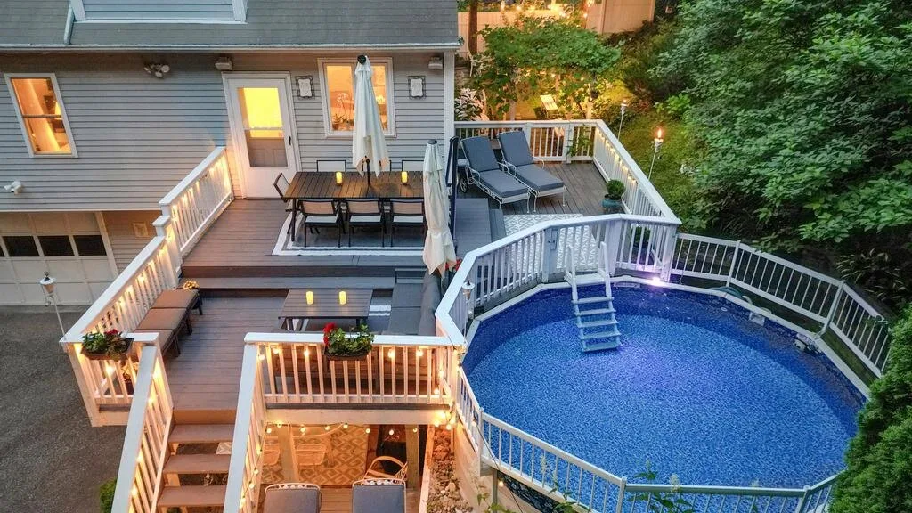 An elevated deck surrounds an above-ground pool beside a house.
