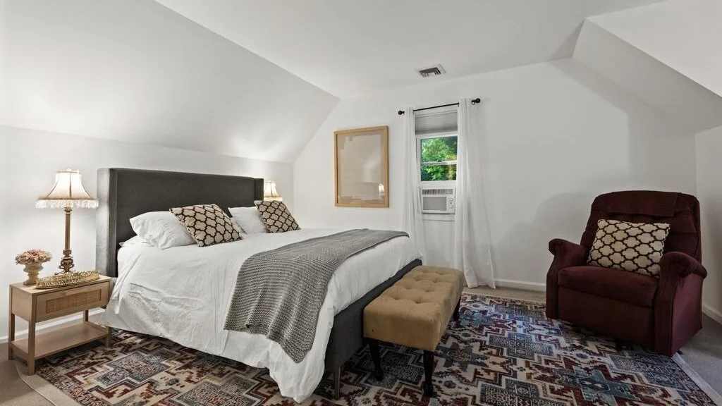 A bedroom with a bed, a burgundy armchair, and patterned carpeting.