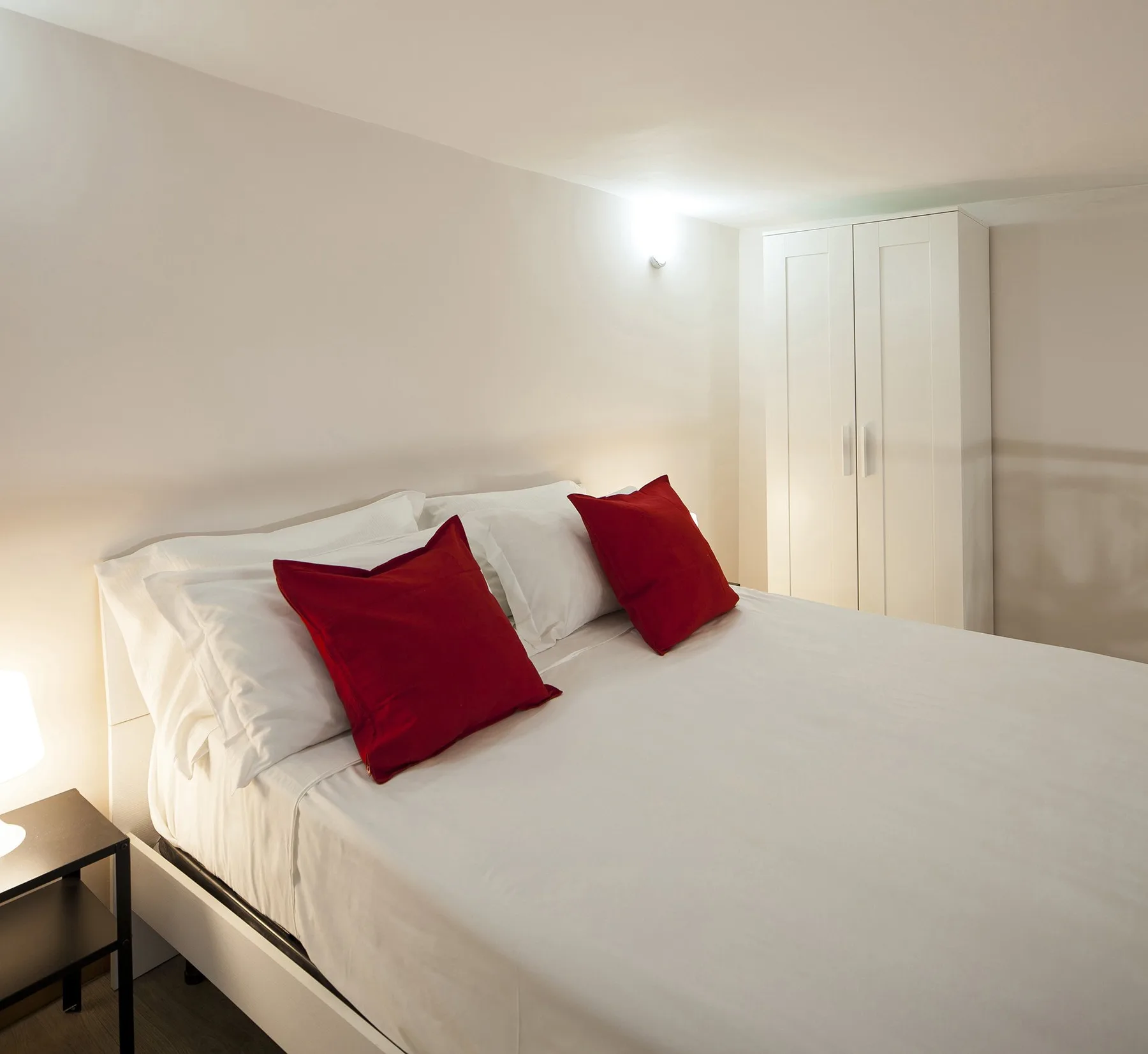 A bedroom with white bedding and red pillows beside a wardrobe.