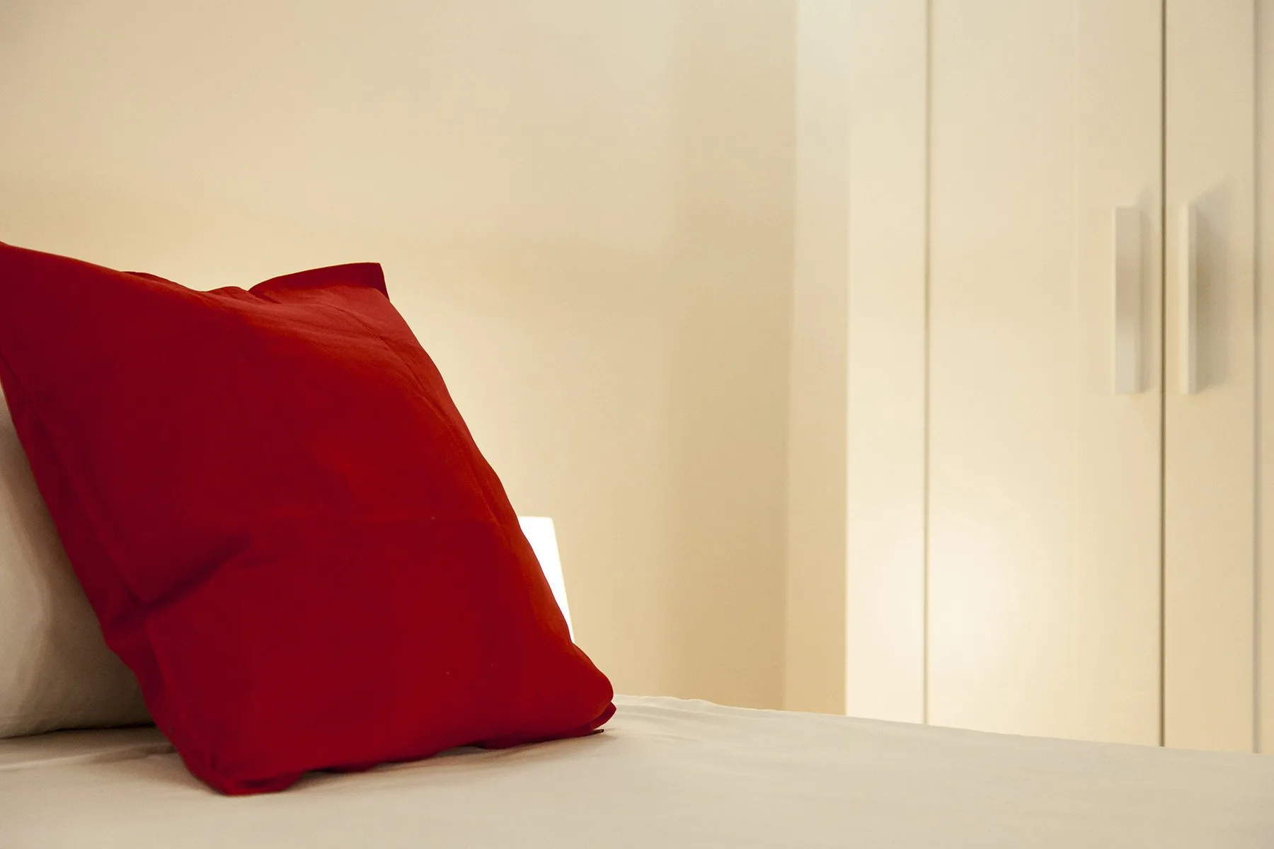 A bed with a bright red pillow sits beside a pale wardrobe.