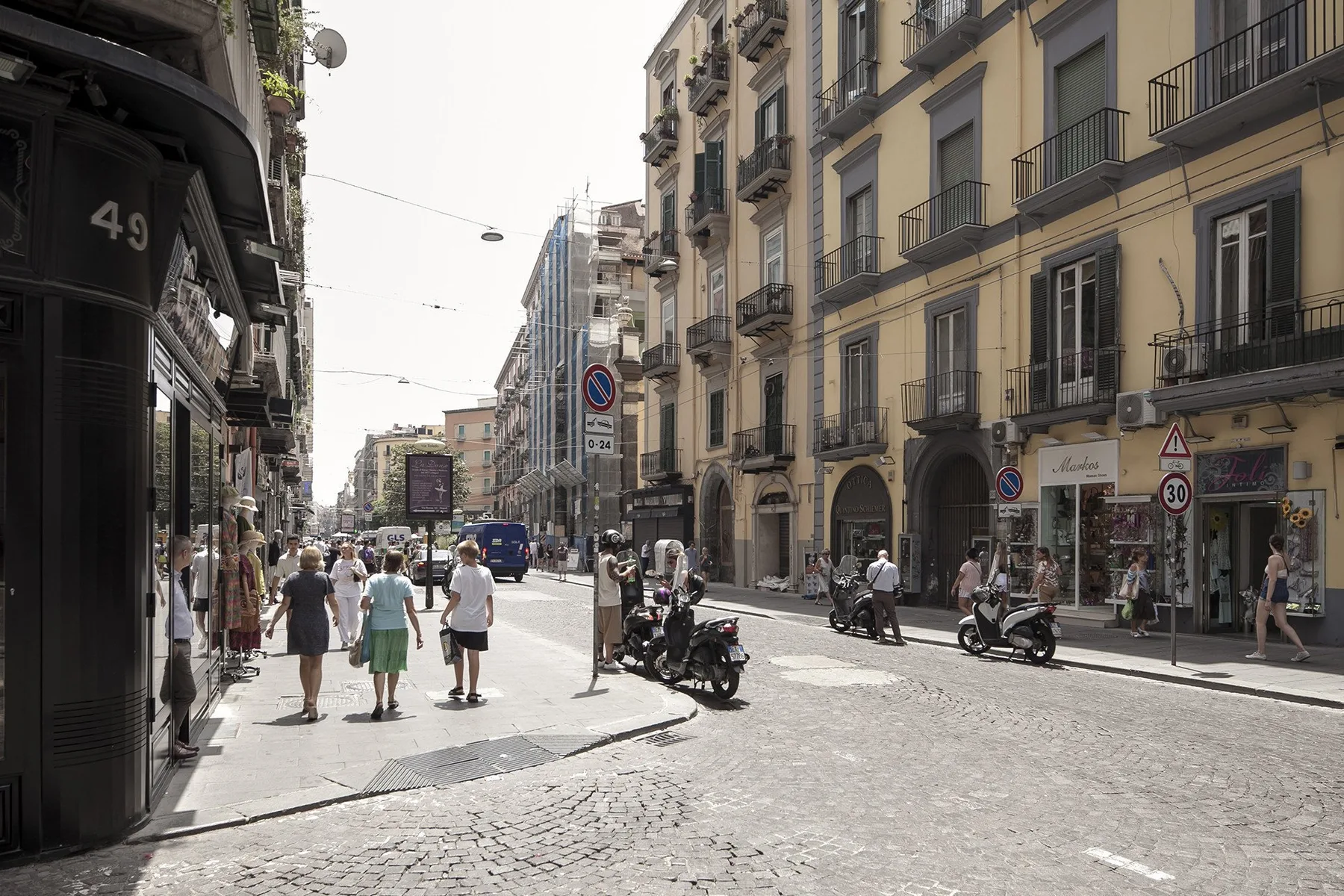 A sunny city street lined with multistory buildings, shops, and parked scooters.