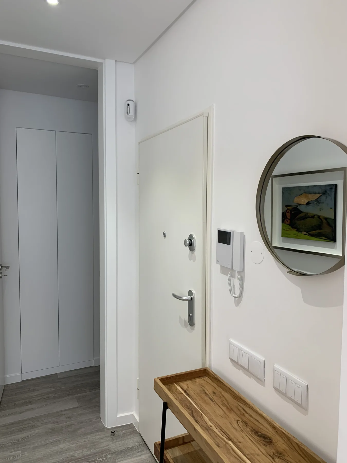 Entrada de un apartamento con puerta blanca, portero de pared y mesa estrecha de madera.