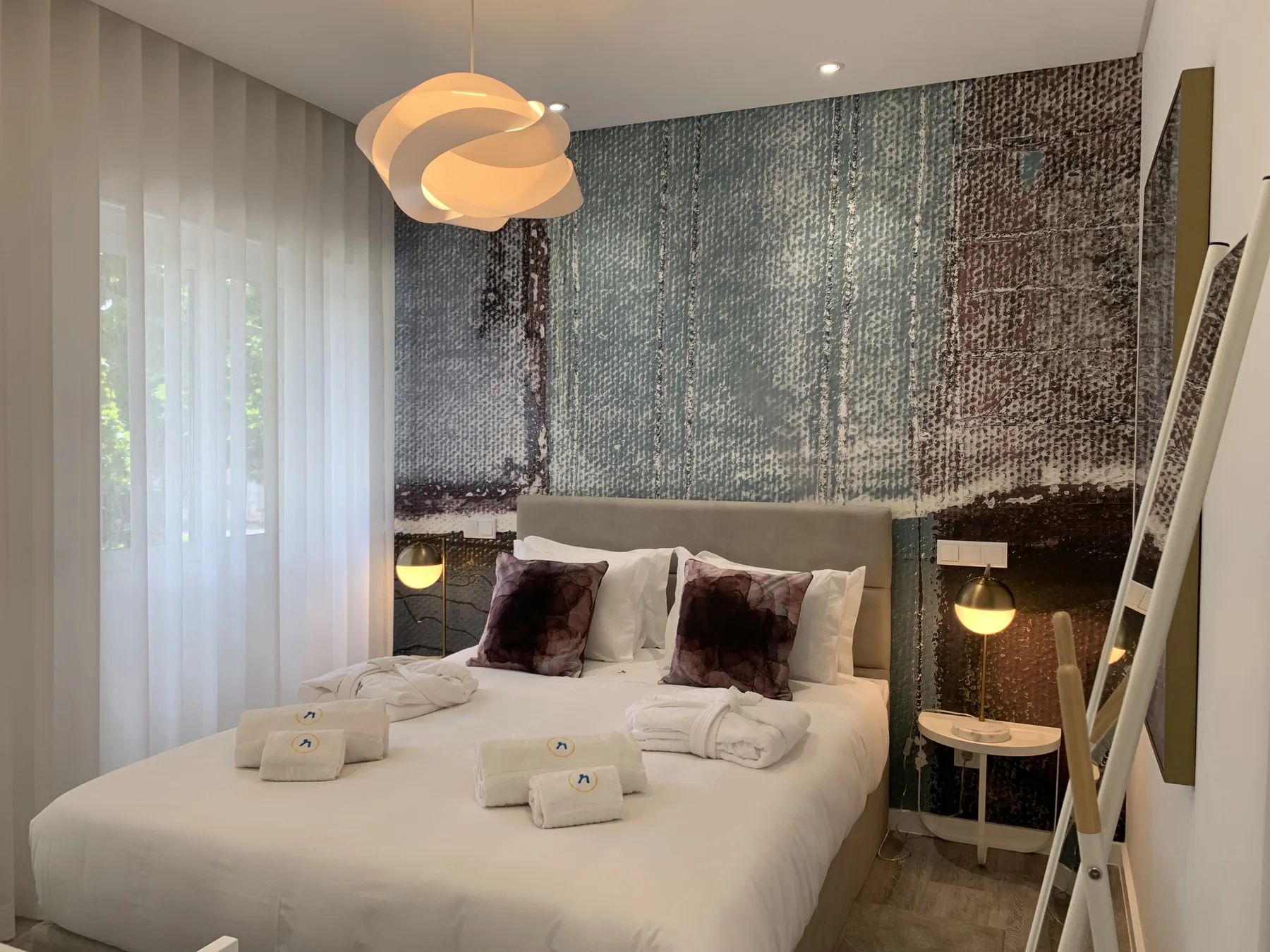 Un dormitorio con una cama bien hecha, una pared estampada y cortinas translúcidas.