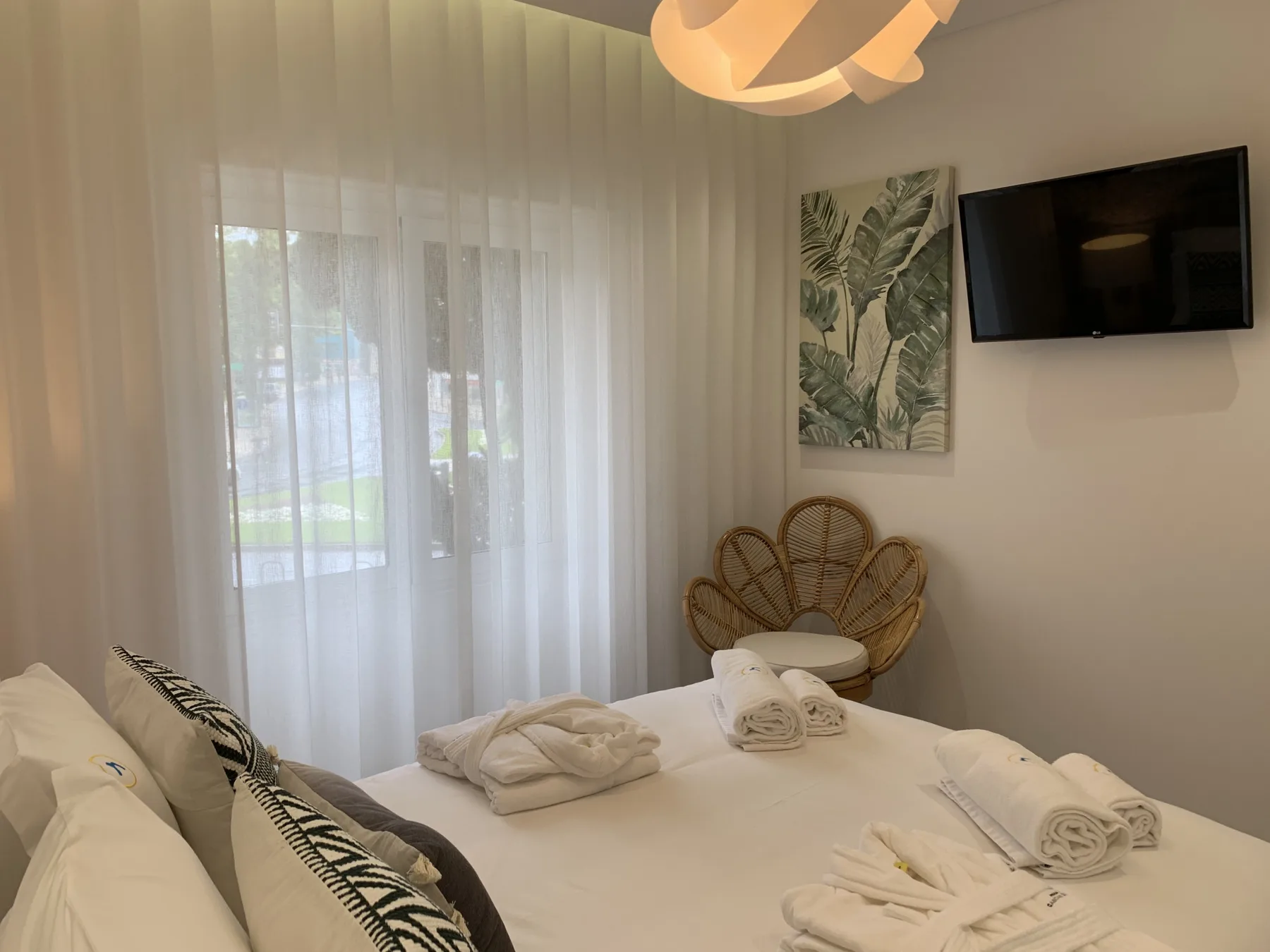 Un dormitorio con una cama ordenada, cortinas translúcidas y un televisor en la pared.
