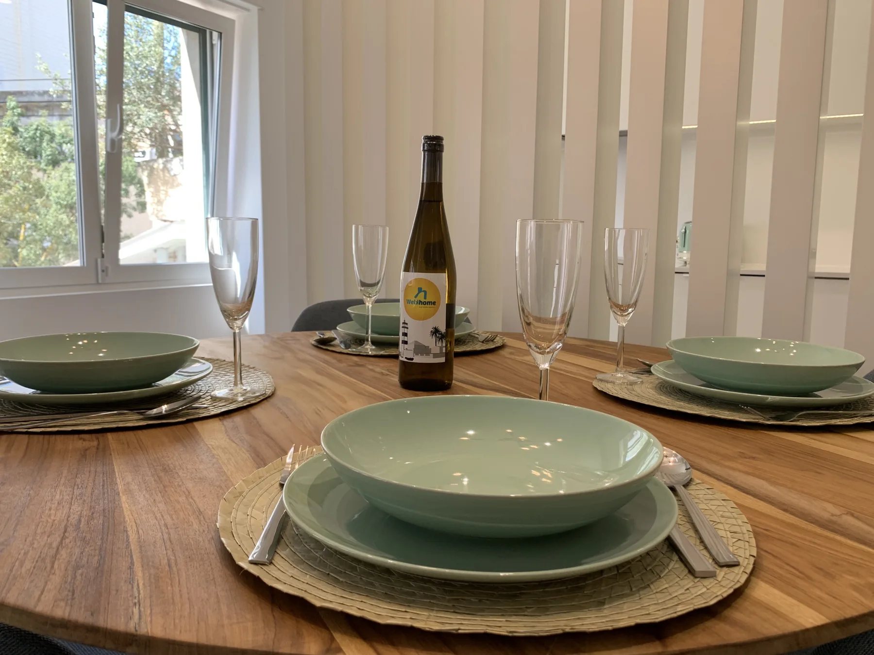 Una mesa de comedor está puesta con vajilla verde y copas junto a una ventana.