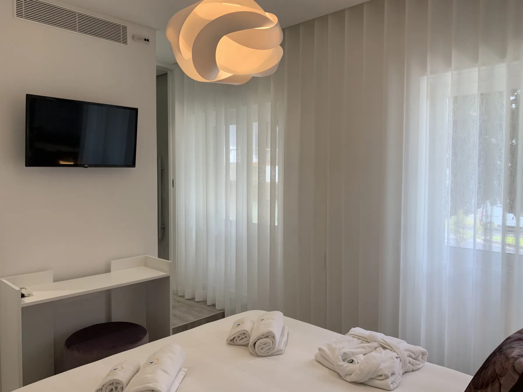 Dormitorio con una cama, cortinas translúcidas y un televisor en la pared.