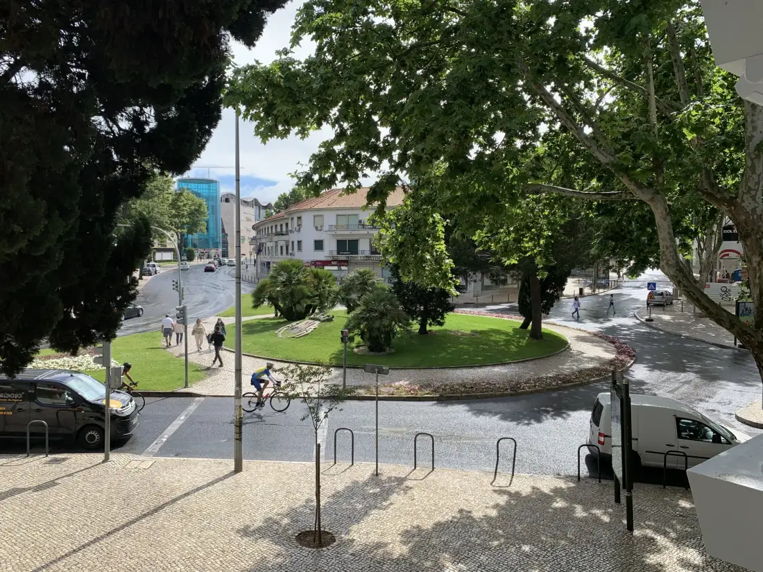 Una rotonda arbolada con una pequeña zona verde, aceras y edificios cercanos.
