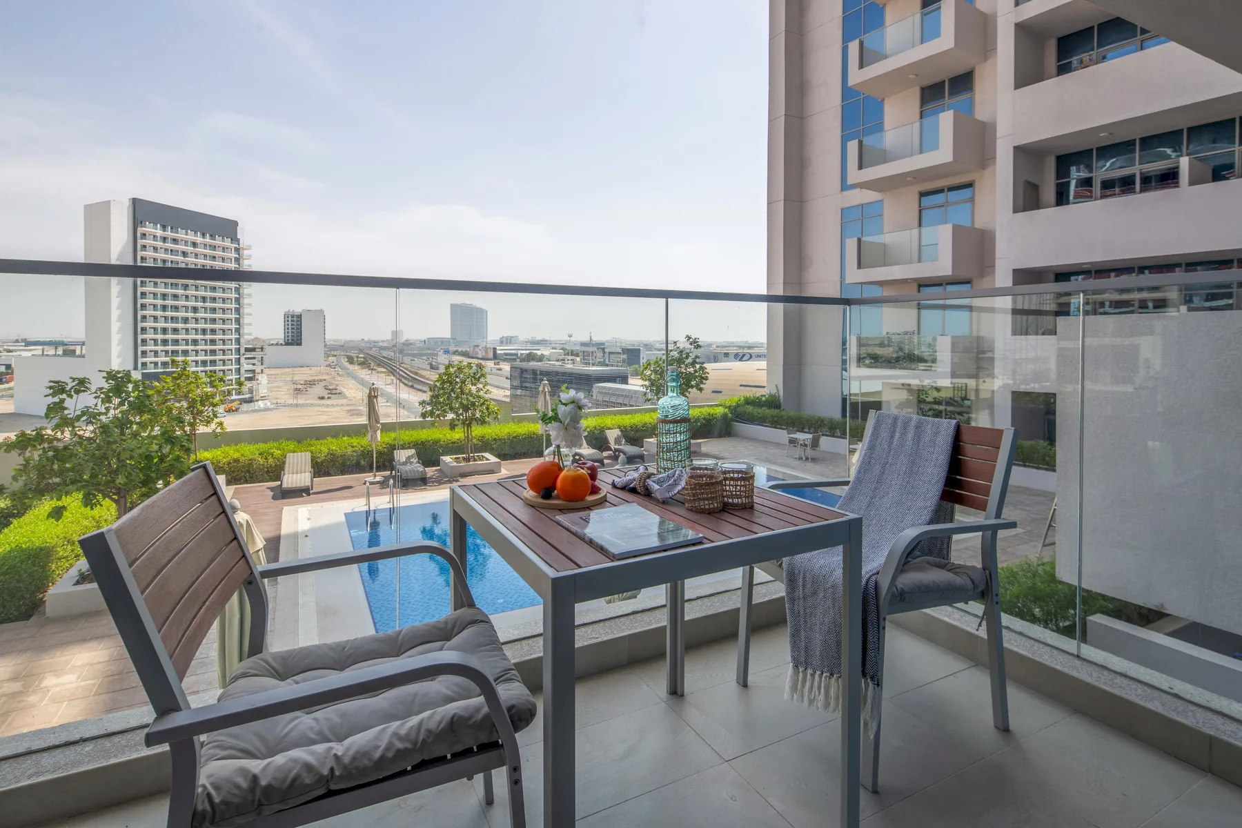 A furnished balcony overlooks a small pool and surrounding city buildings.