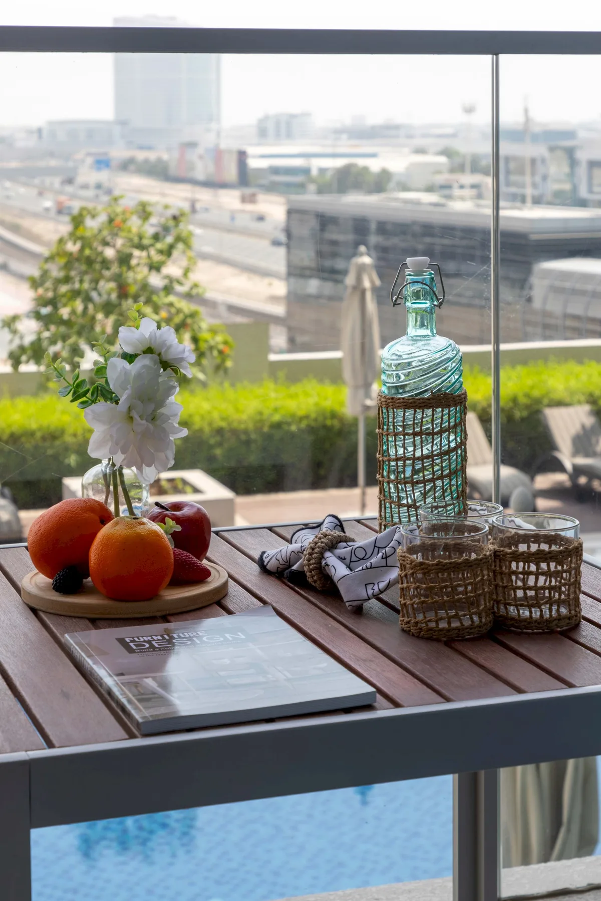 A patio table with flowers and a glass bottle overlooks a swimming pool and city.