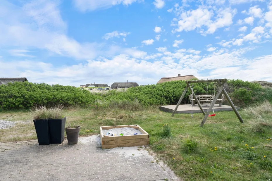 Una zona de juegos al aire libre con un arenero de madera y un columpio junto a dunas cubiertas de hierba.
