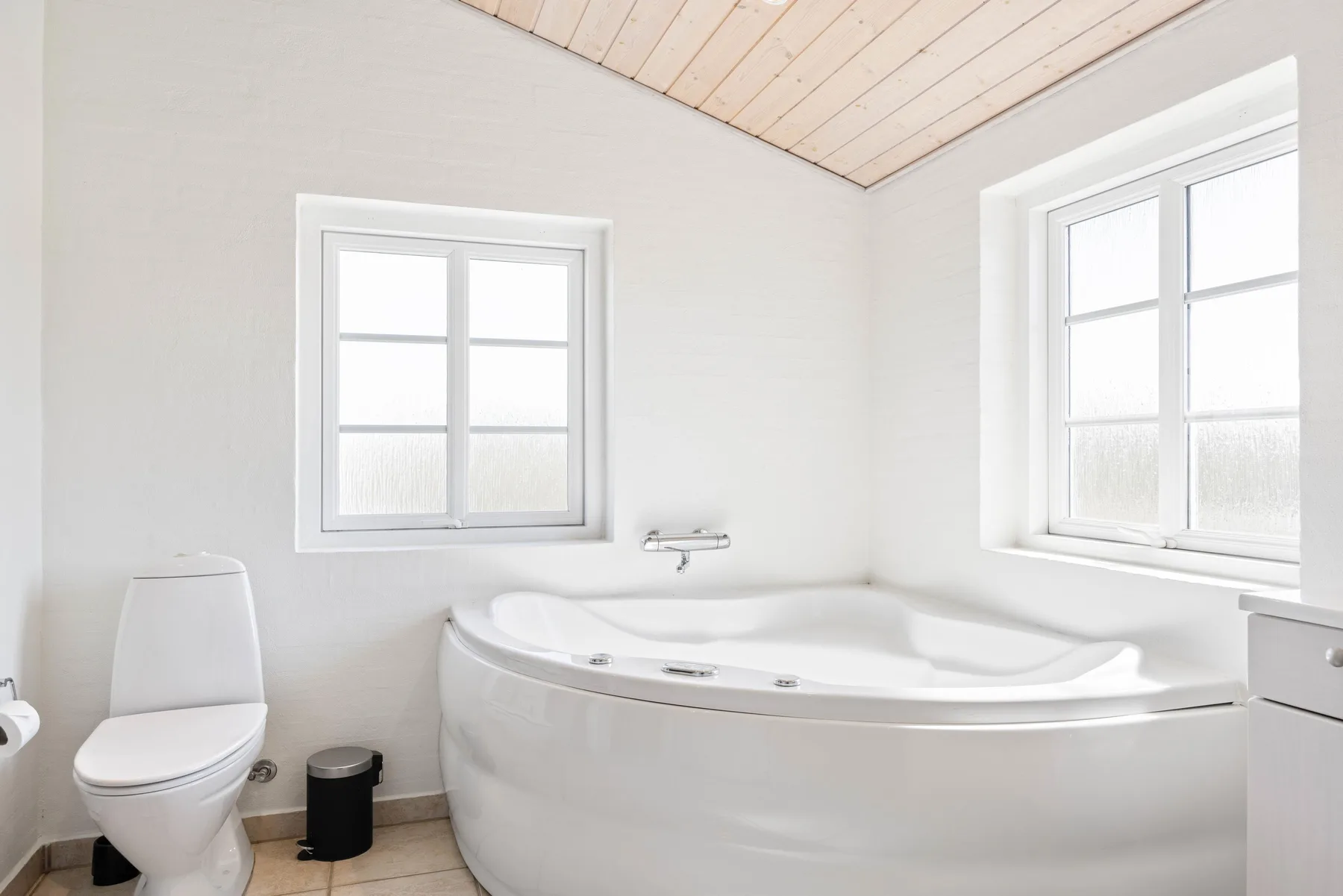 Un baño luminoso con una bañera grande bajo dos ventanas.