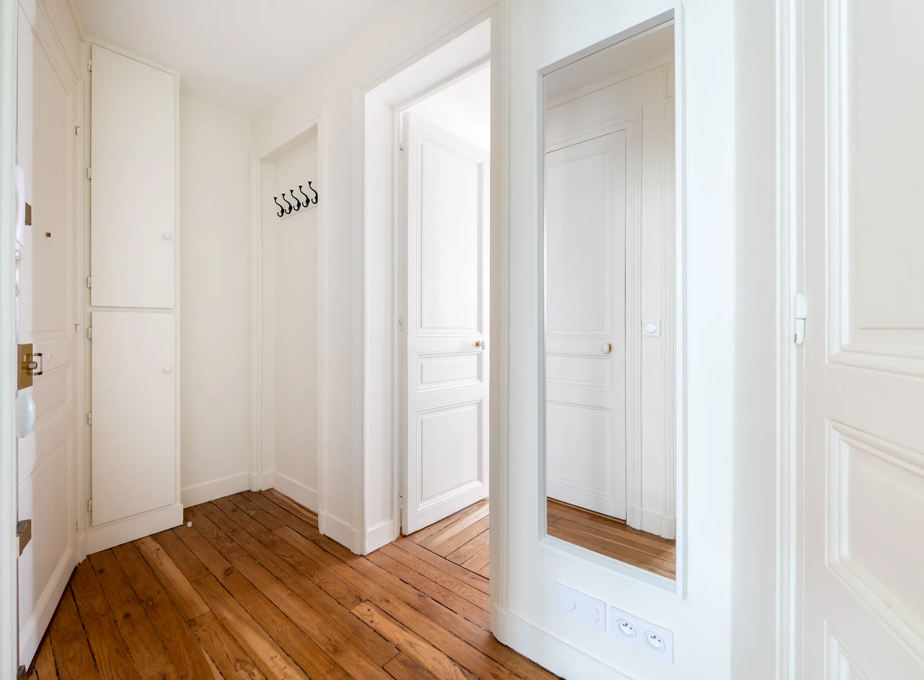 A bright hallway with wooden flooring, white doors, and a wall-mounted coat rack.