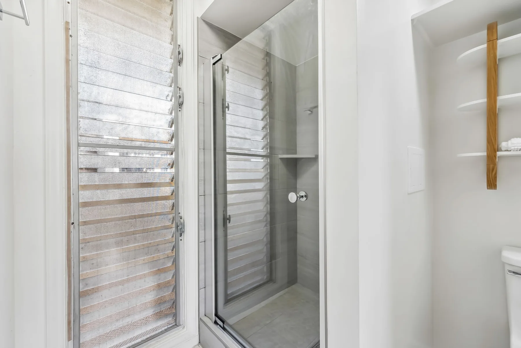 Un baño luminoso con una ducha de vidrio y una ventana con persianas horizontales.