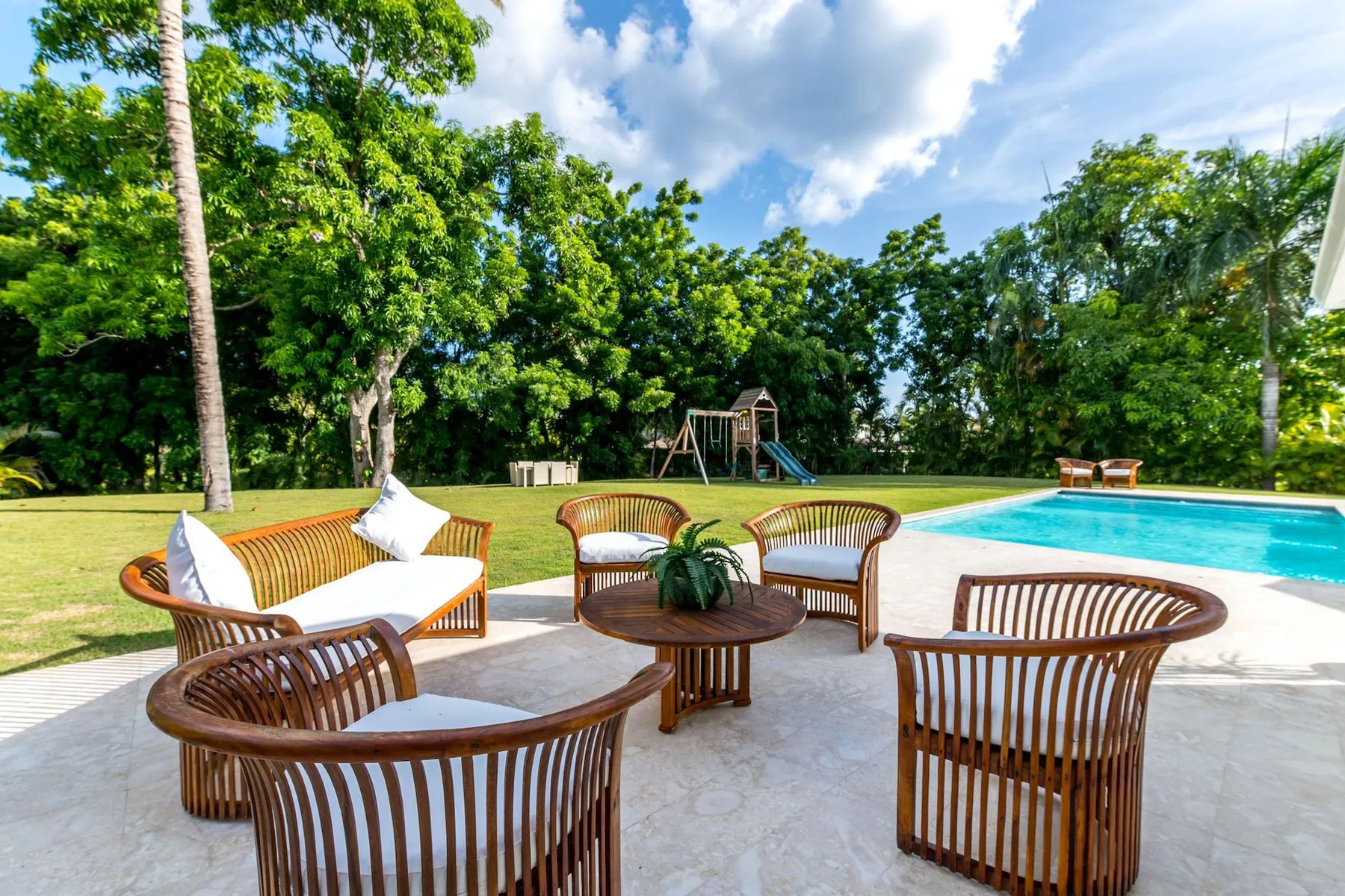 An outdoor pool patio has wooden seating and a tree-lined lawn.