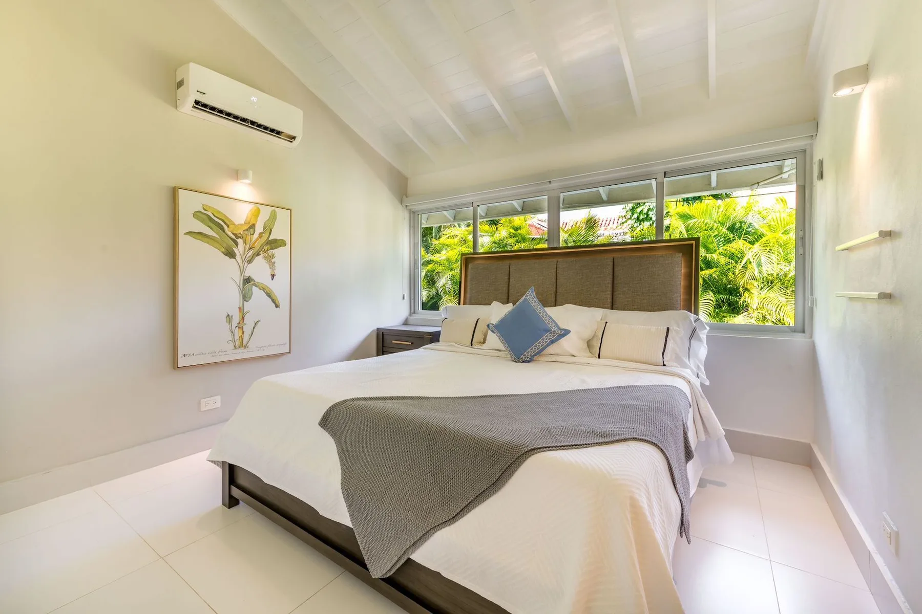 A bright bedroom with a large bed and windows overlooking green foliage.