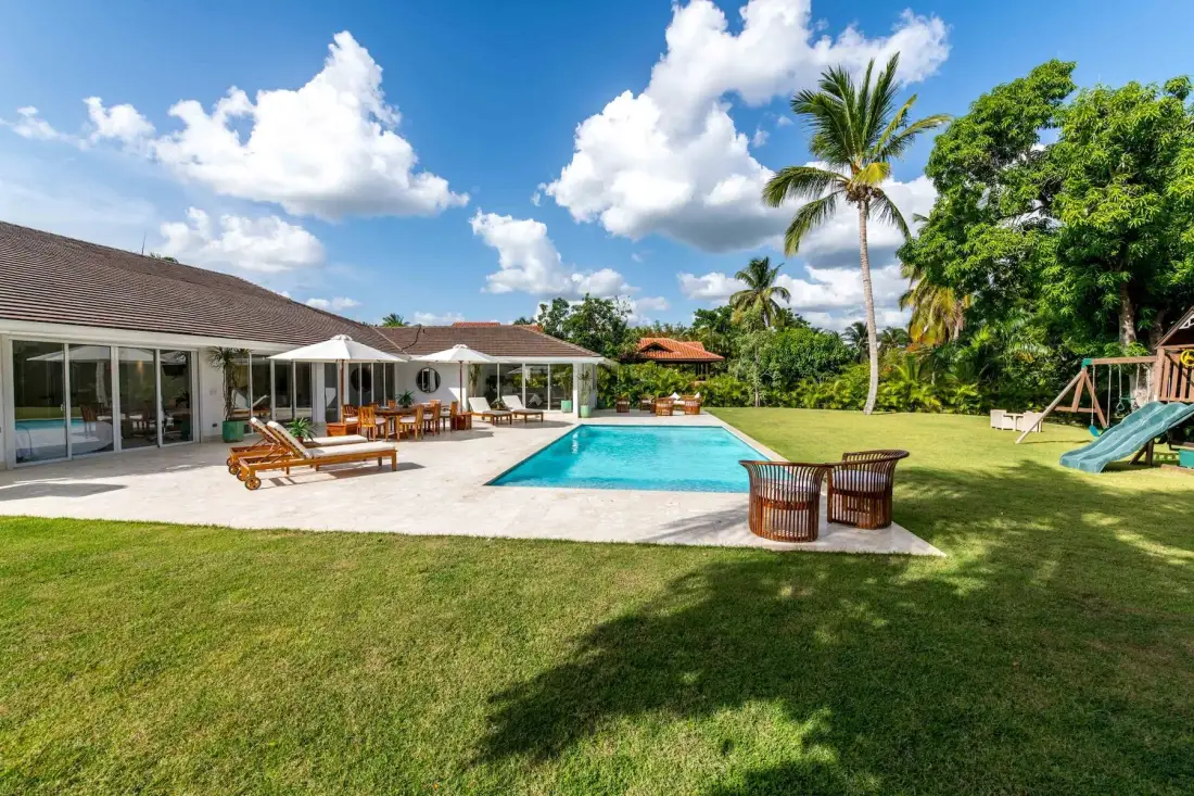 A swimming pool sits beside a patio, lawn, and low white buildings.