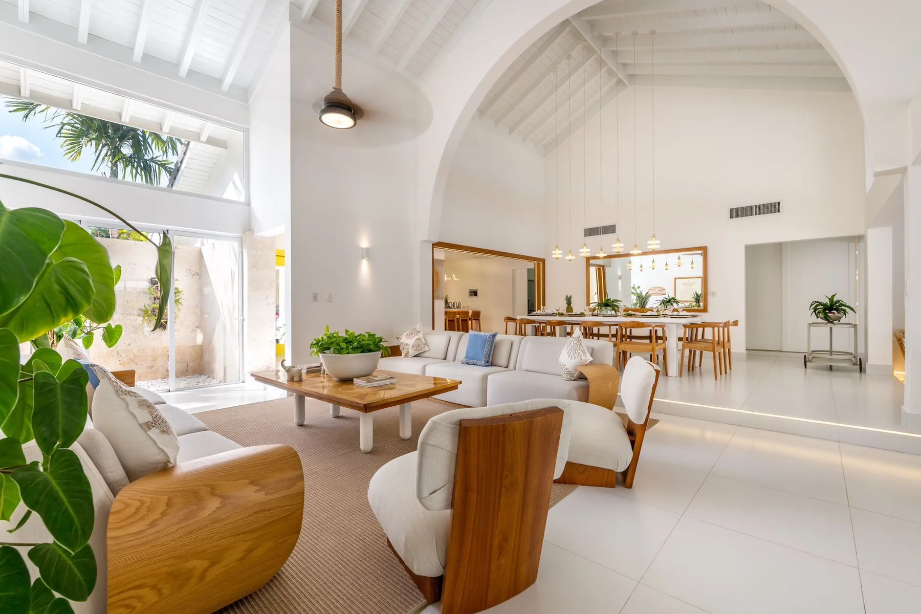 A bright living and dining area features seating, a dining table, and large leafy plants.