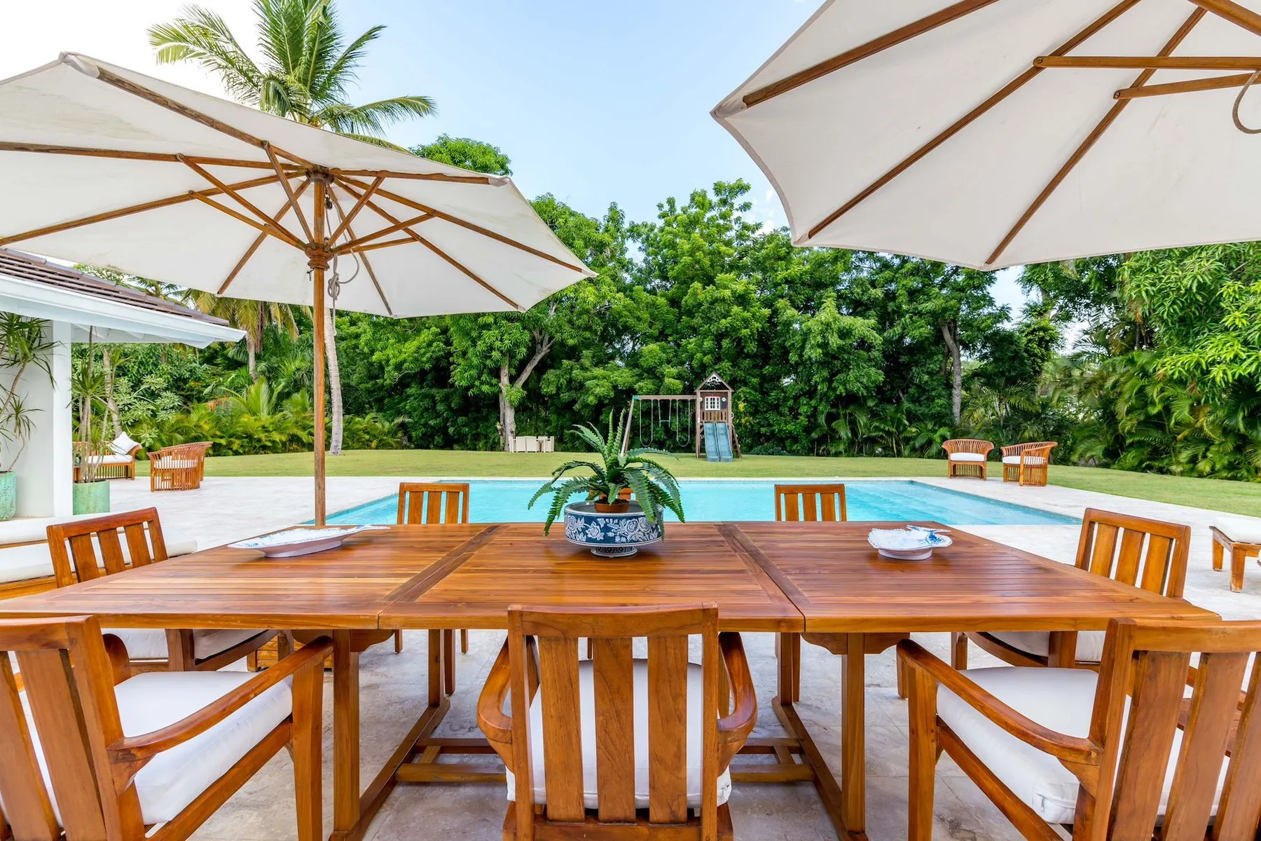 An outdoor dining area beside a swimming pool, shaded by large umbrellas.