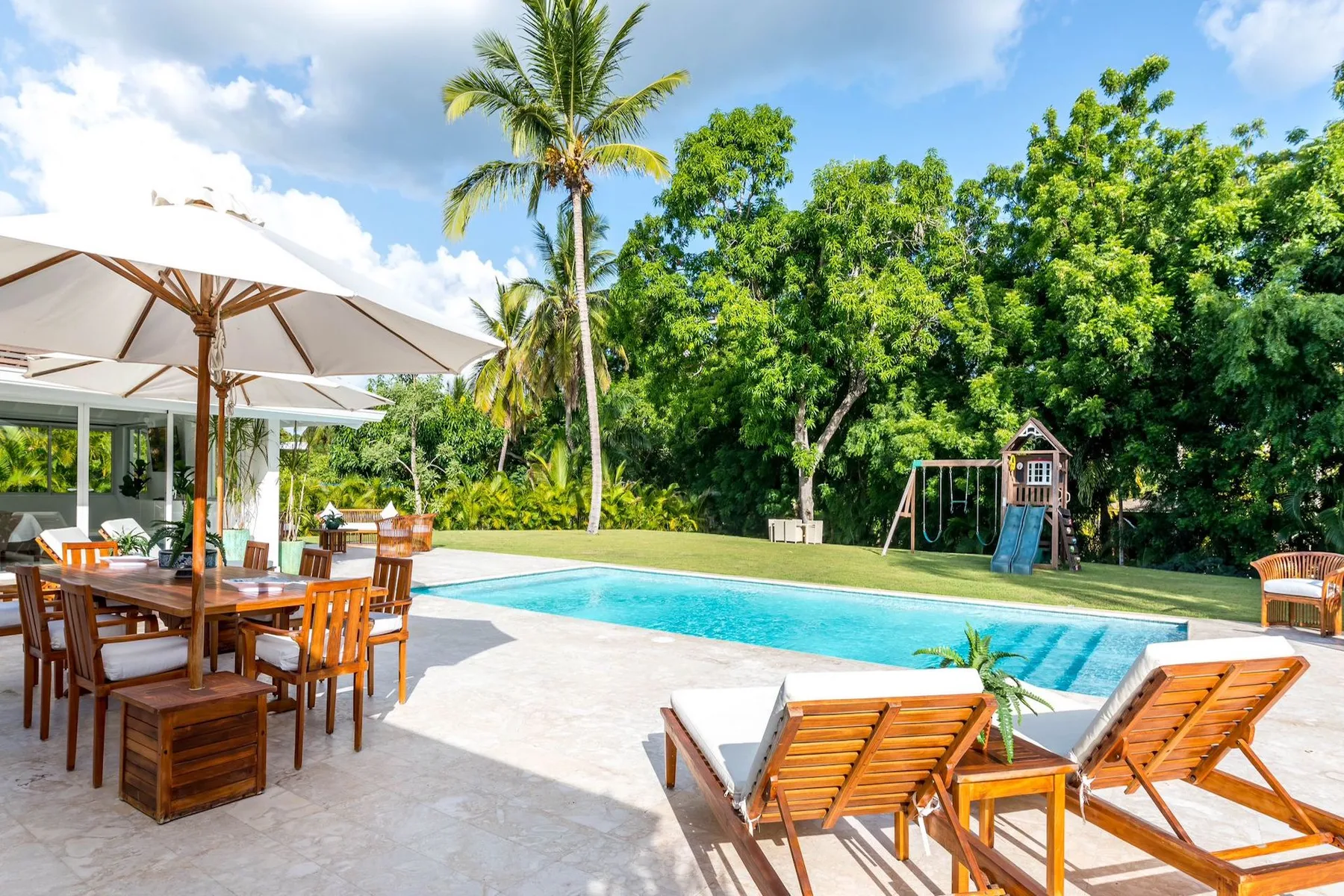 An outdoor pool area has wooden seating and a shaded dining space among tropical greenery.