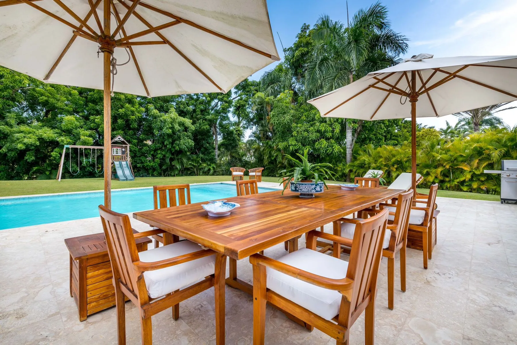 An outdoor dining area with a wooden table and umbrellas beside a swimming pool.