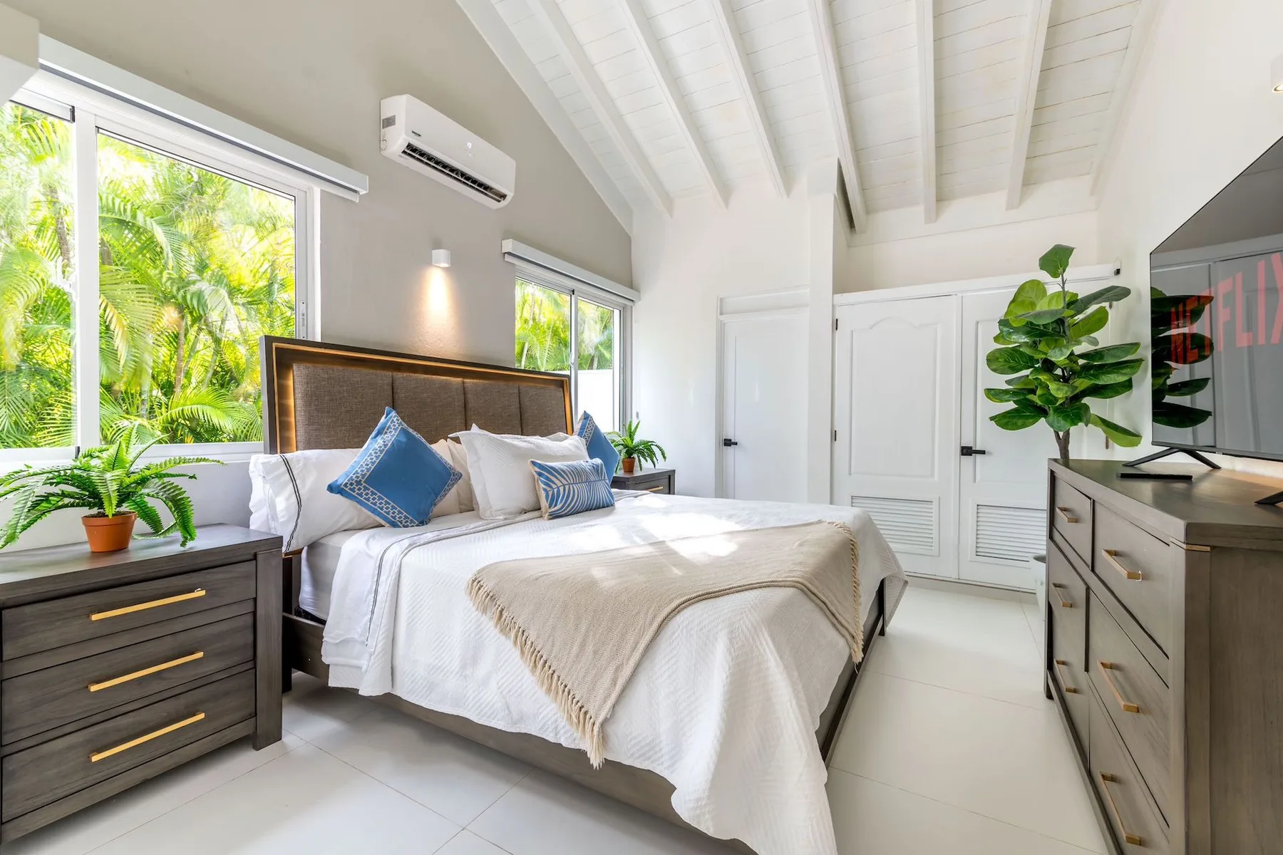 A bright bedroom with a bed, tall windows, and leafy plants.