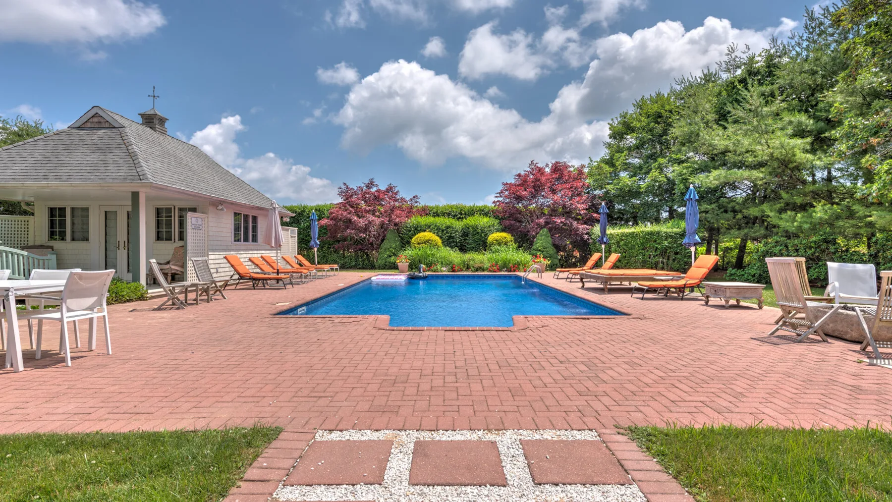 Una piscina al aire libre está en un patio de ladrillo junto a una casa y un jardín.