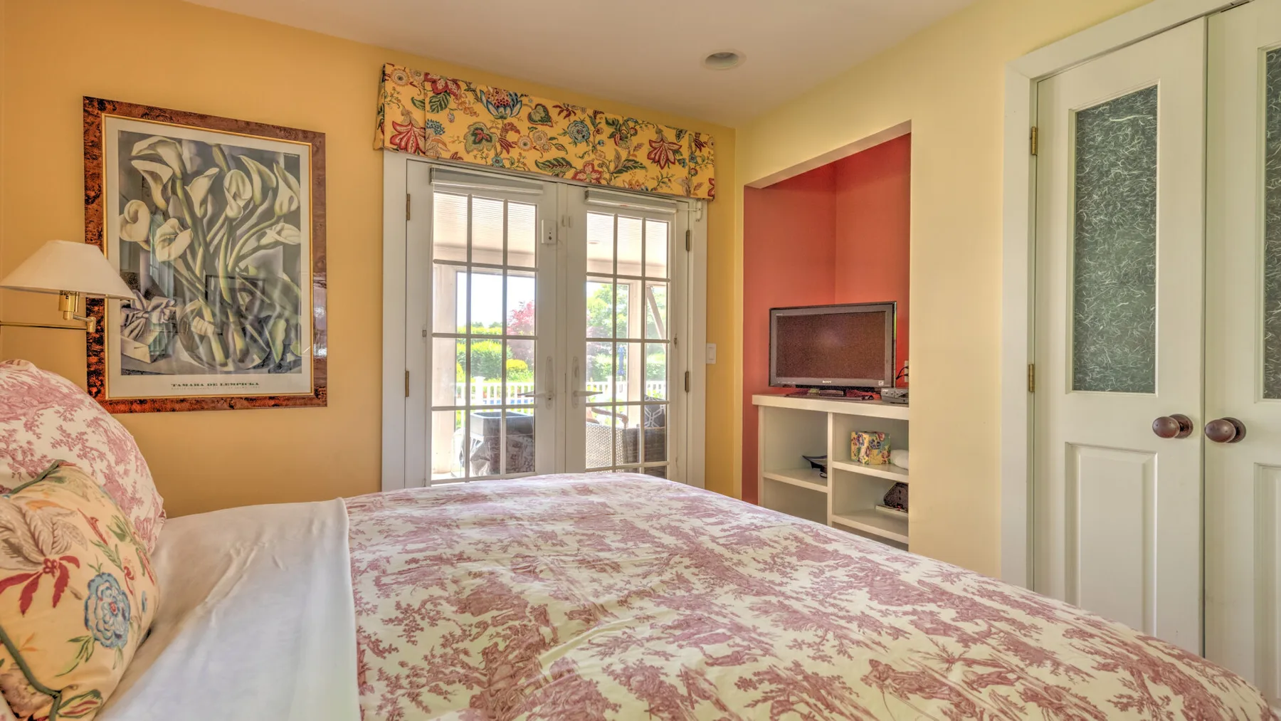 Un dormitorio con una colcha floral, arte enmarcado y puertas de vidrio que dan a un patio.