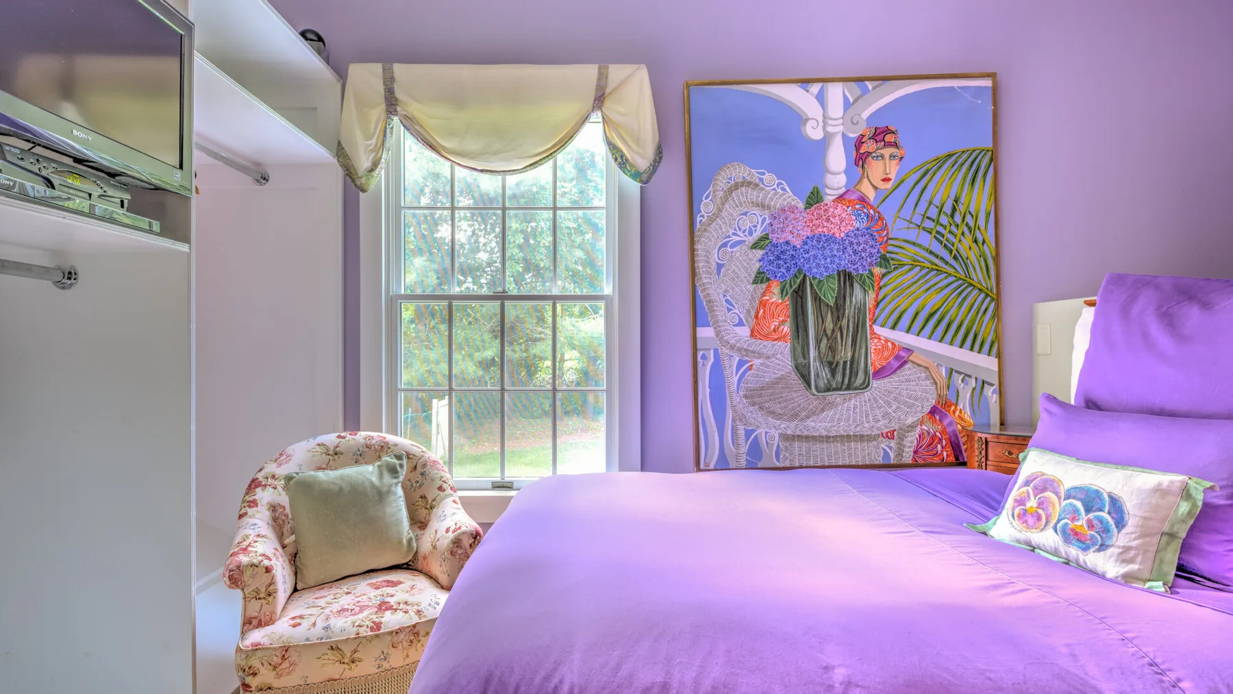 Un dormitorio con una cama morada, un sillón floral y una ventana grande.