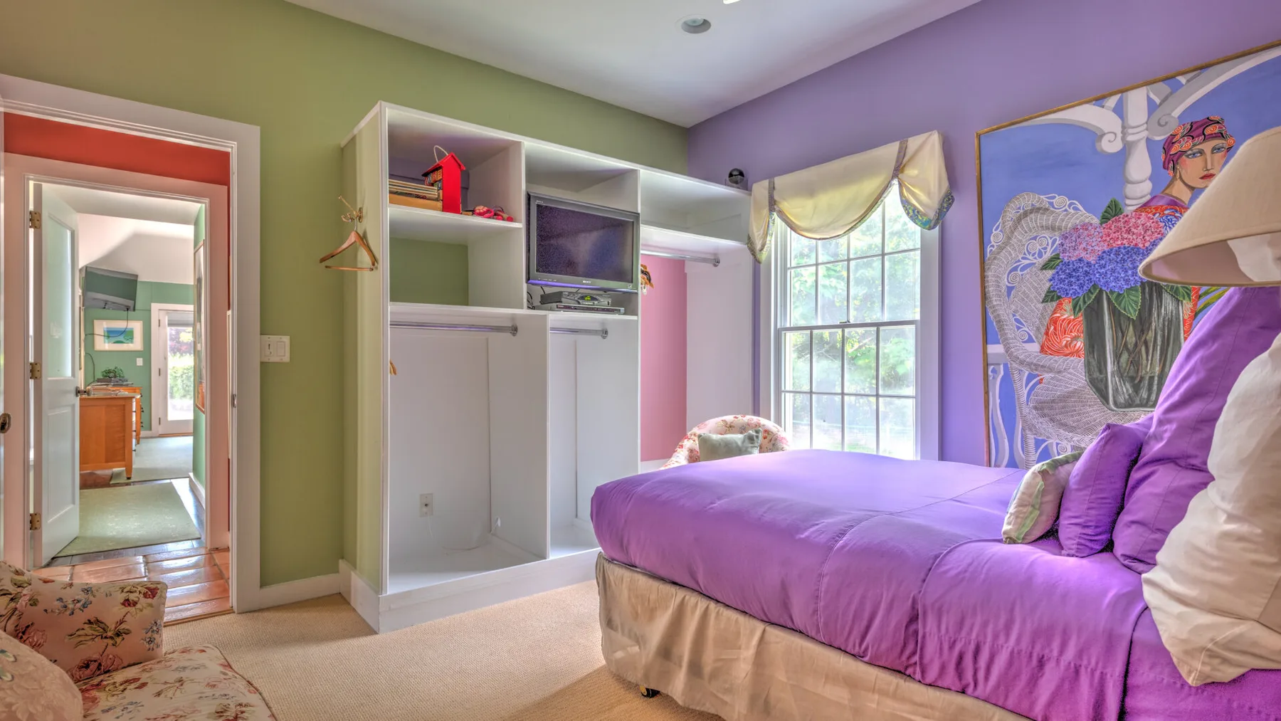 Un dormitorio con una cama morada junto a una ventana grande y arte colorido en la pared.