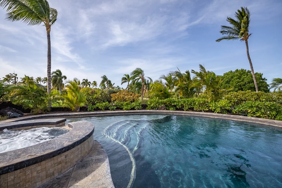 Una piscina exterior curva está junto a un jacuzzi burbujeante, con palmeras al fondo.