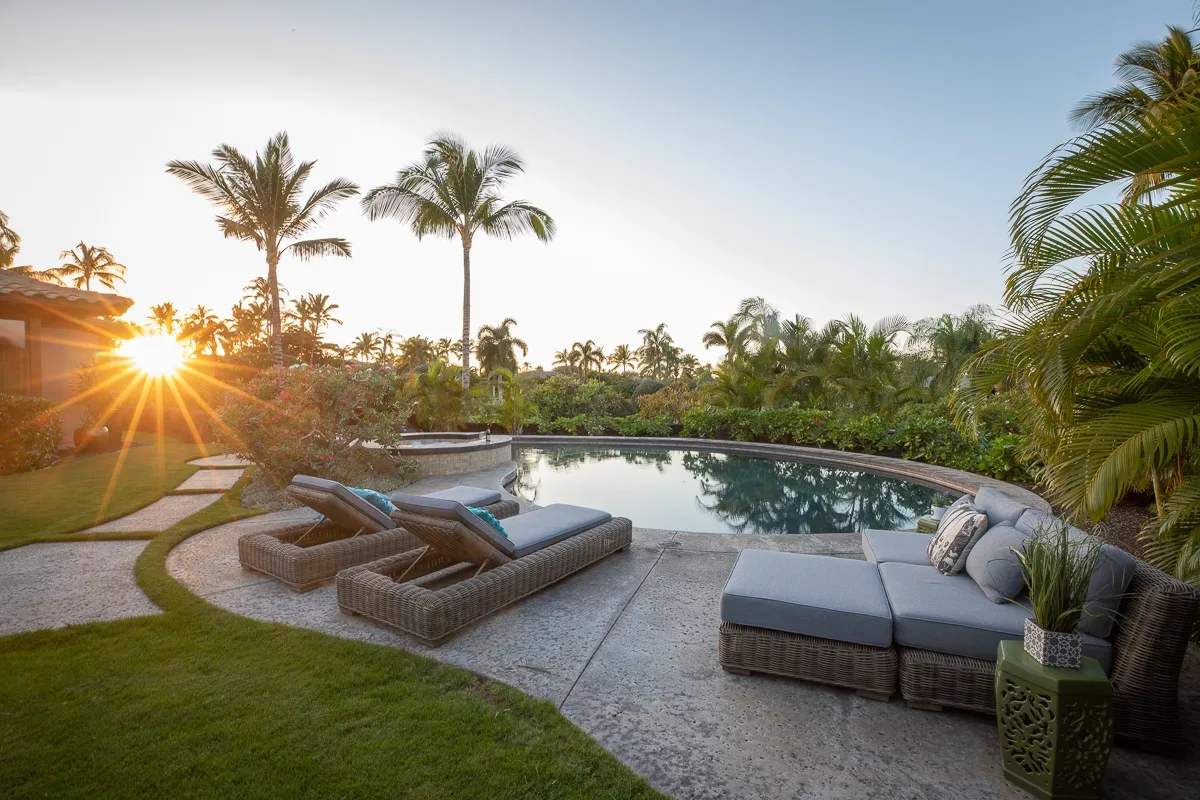 Una piscina y asientos en el patio están rodeados de plantas tropicales al atardecer.