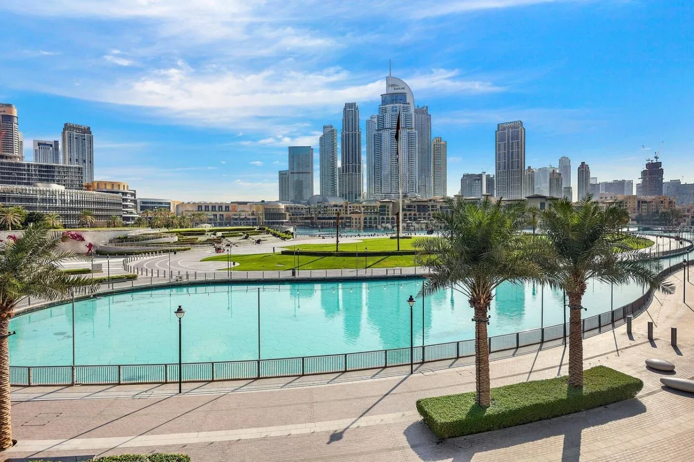 Un paseo curvo junto al agua bordea una piscina turquesa, con palmeras y un perfil urbano al fondo.