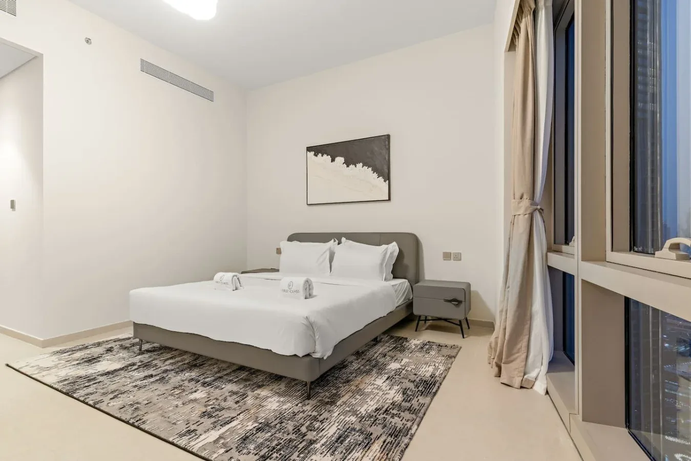 Un dormitorio con una cama bien tendida, una mesa de noche y ventanas grandes.