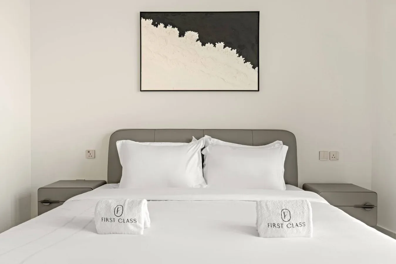 Una cama bien tendida con ropa de cama blanca está debajo de una lámina abstracta enmarcada.