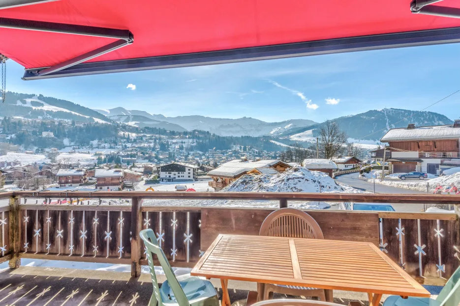 A balcony dining area overlooks a snowy mountain town beneath a red awning.