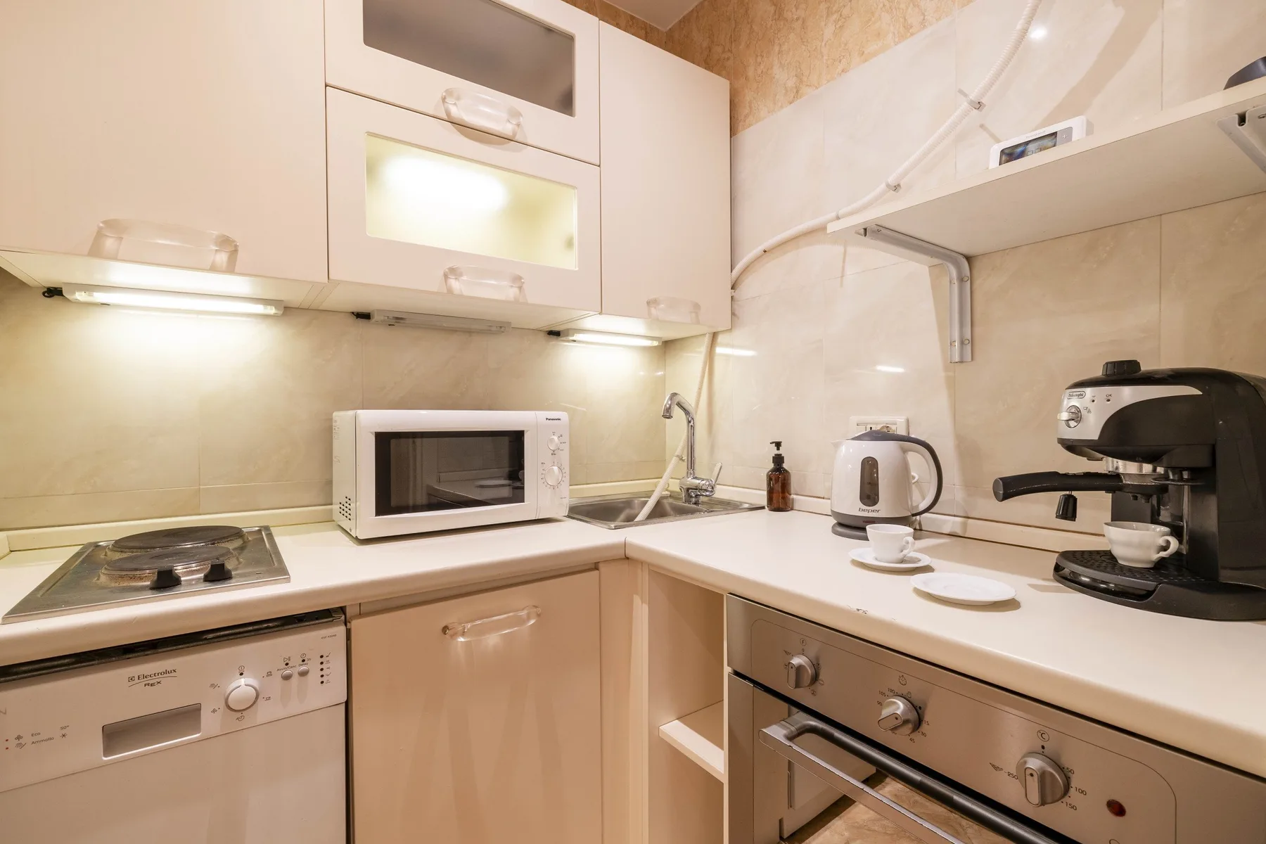 A compact kitchen with a cooktop, microwave, kettle, and coffee machine on the counters.