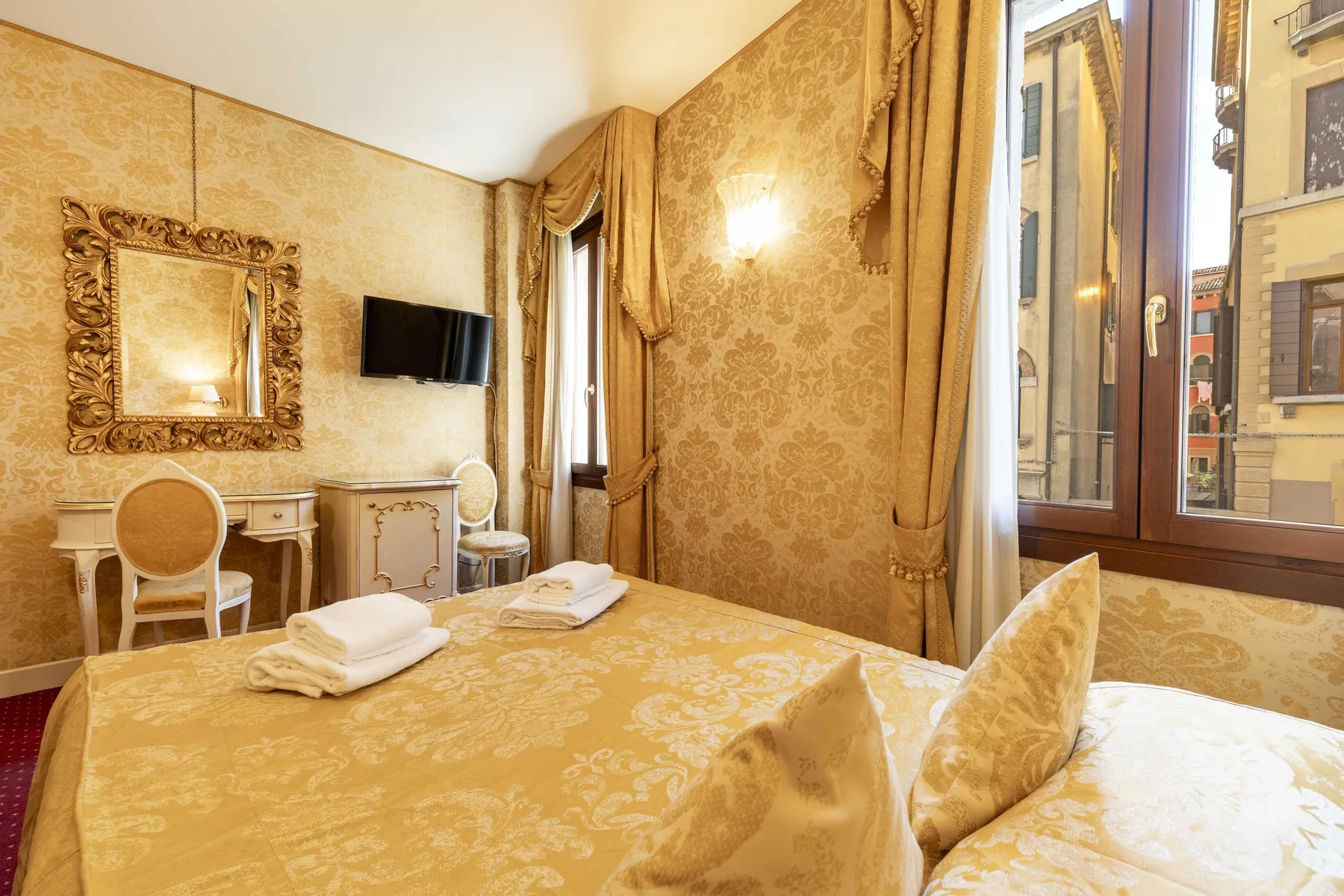 A bedroom with gold-patterned bedding and a large window overlooking nearby buildings.