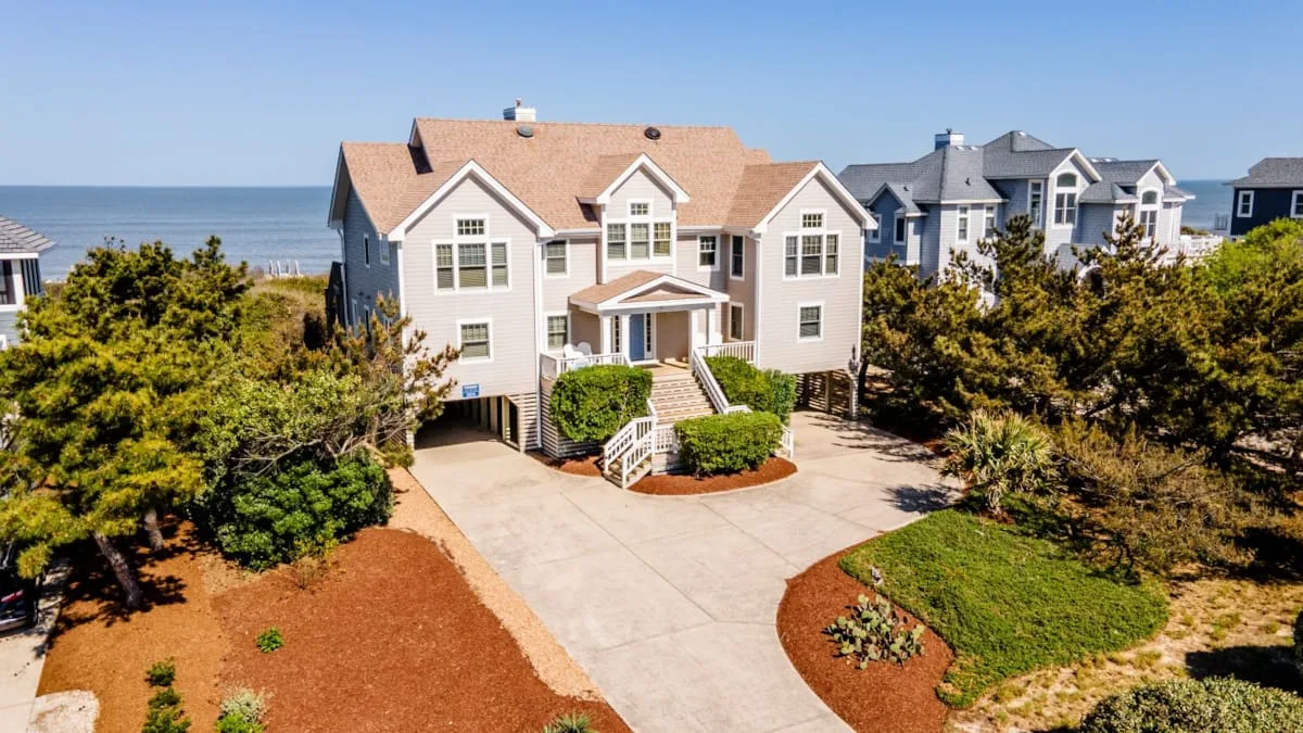 A large coastal house stands beside a paved driveway and landscaped greenery.