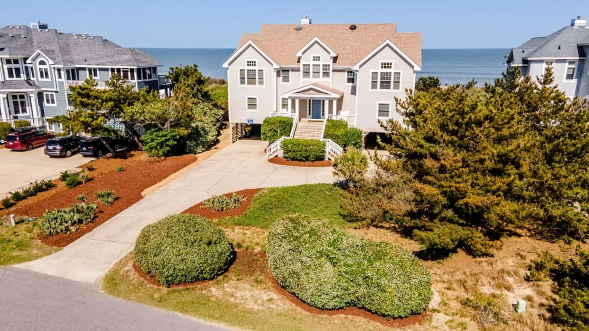 A coastal residential building stands beside a landscaped driveway, with the ocean visible behind it.