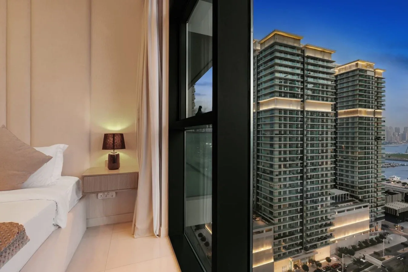 Dormitorio con una mesita de noche junto a grandes ventanas con vistas a edificios altos.