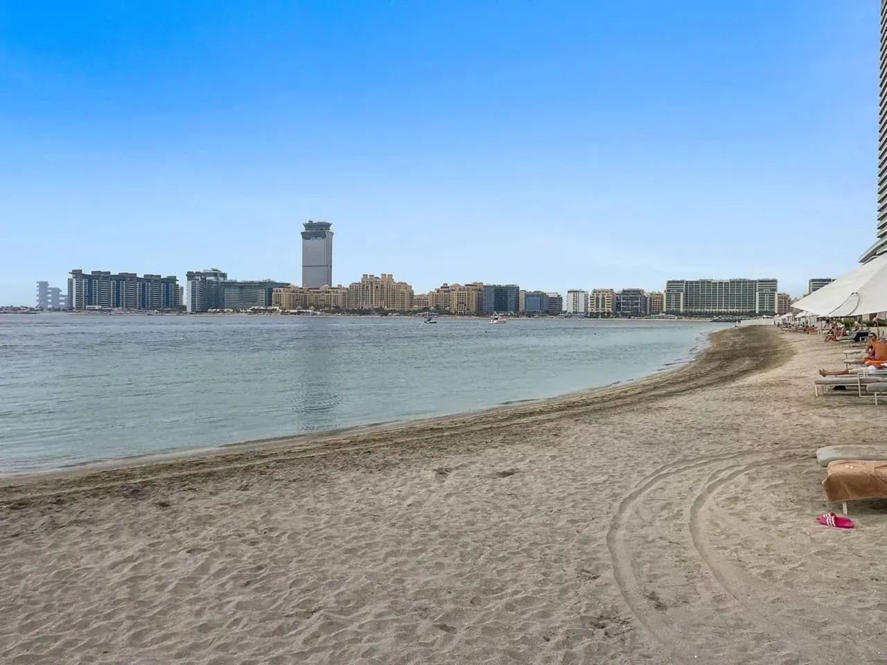 Una playa de arena bordea aguas tranquilas, con edificios visibles al otro lado de la bahía.
