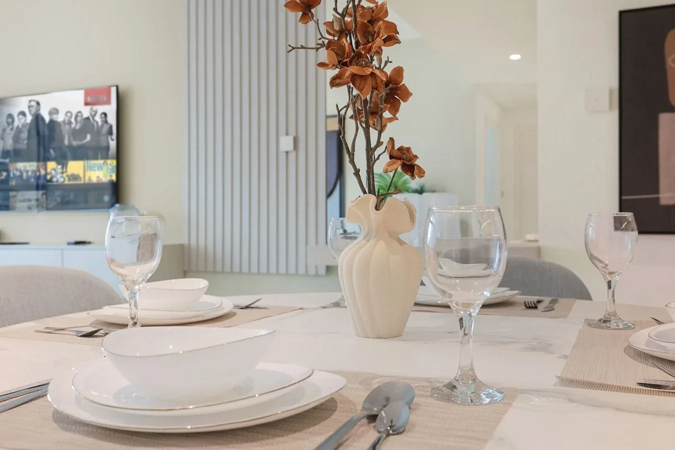 Una mesa de comedor puesta con platos y copas está junto a un jarrón con flores secas.