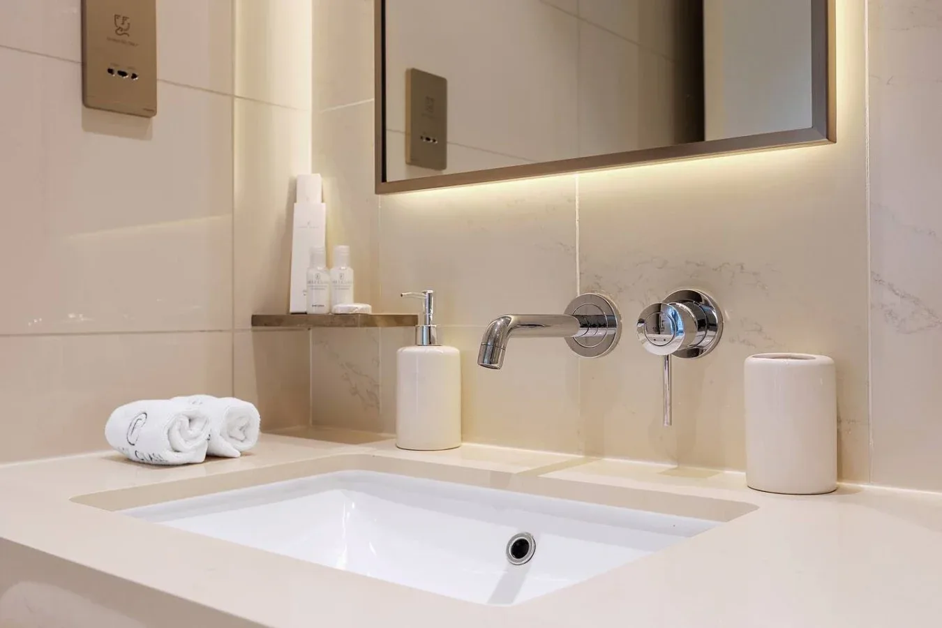 Un lavabo de baño con fregadero blanco, grifo de pared y espejo iluminado.