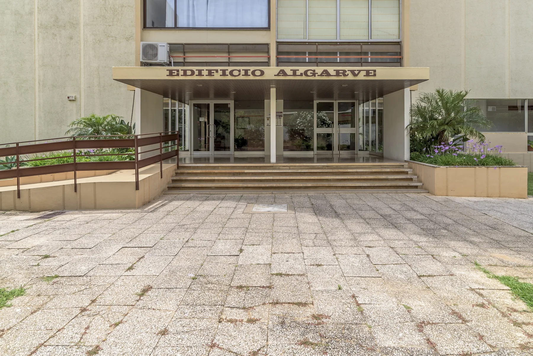 Entrada del edificio con puertas de vidrio, escalones amplios y patio pavimentado.