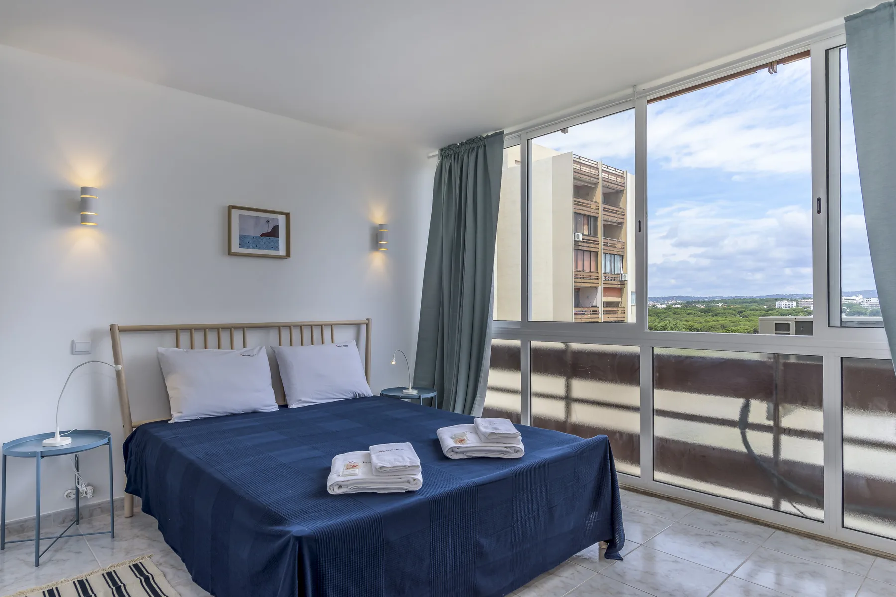 Un dormitorio con una cama junto a grandes ventanas con vistas a edificios y vegetación.