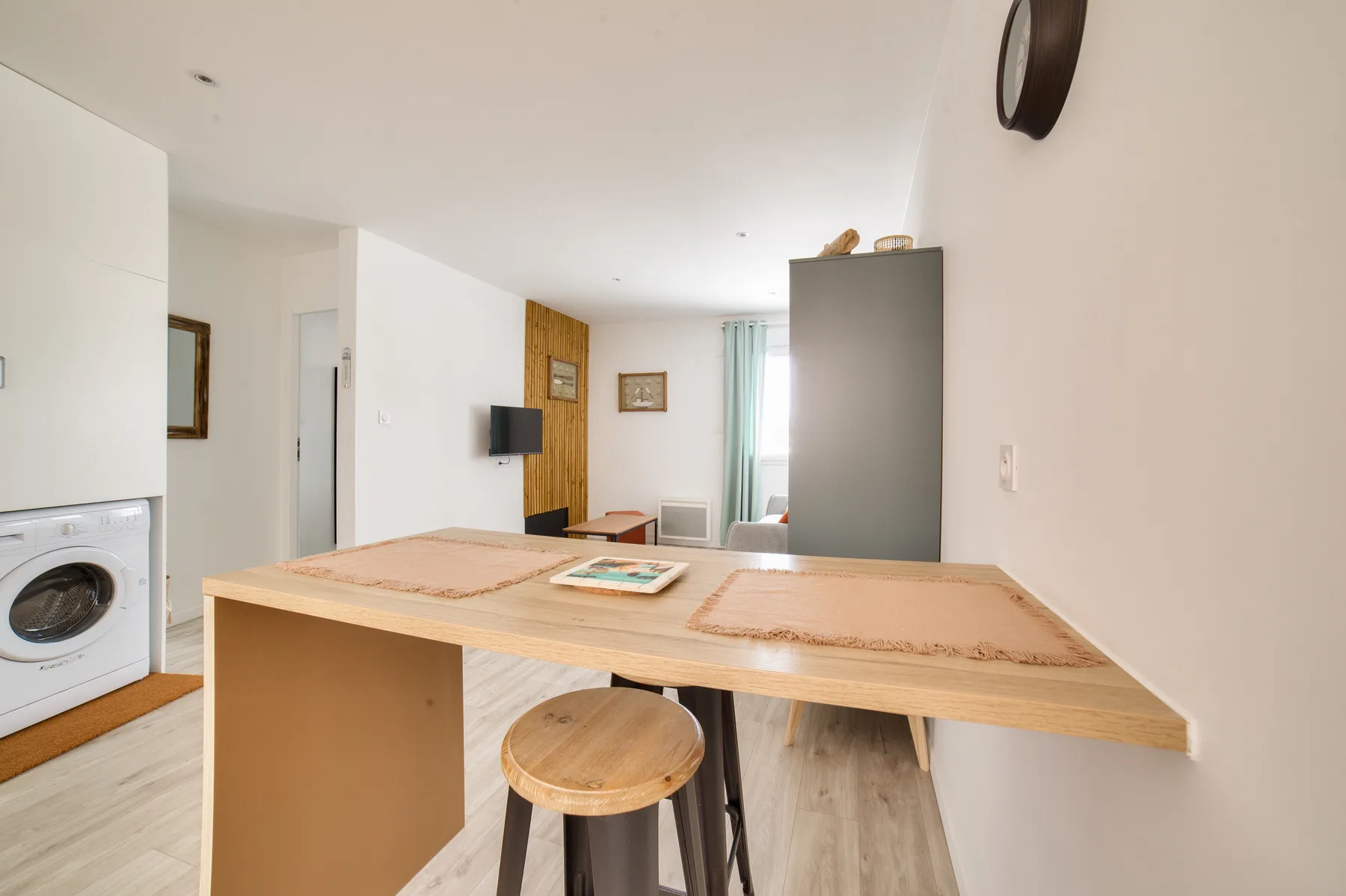 A compact studio interior with a wooden counter, stools, and a washing machine.