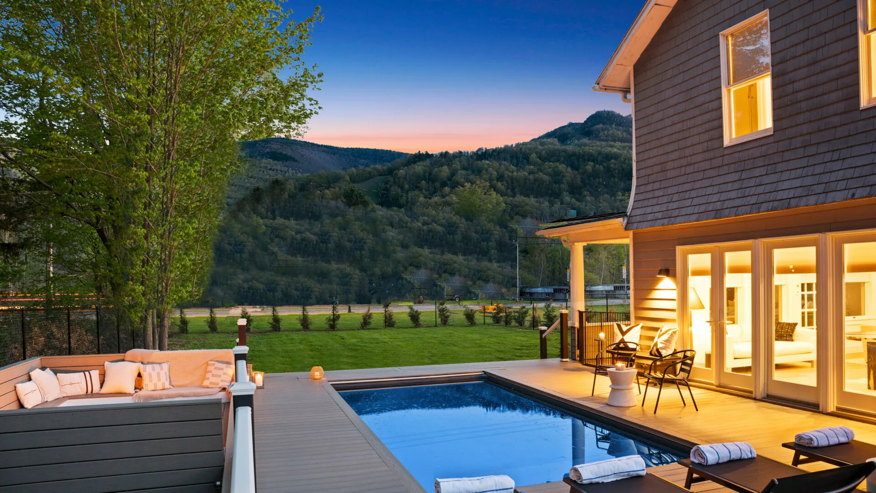 A modern house and outdoor pool overlook a green lawn and wooded hills at dusk.