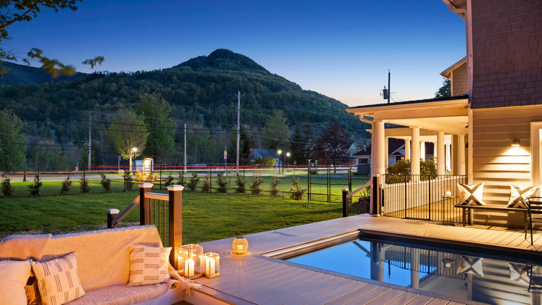 A patio beside a swimming pool overlooks a green lawn and wooded hills at dusk.