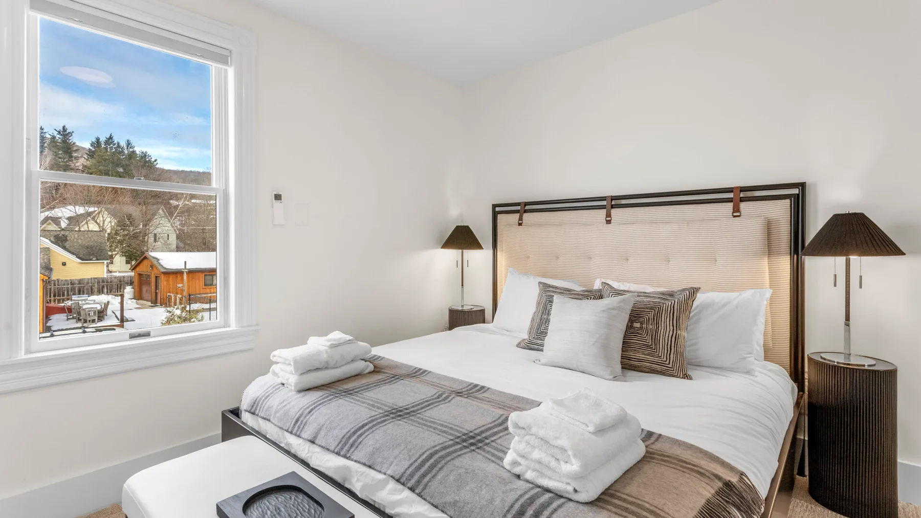 A bright bedroom with neatly made bedding and a window overlooking houses and trees.
