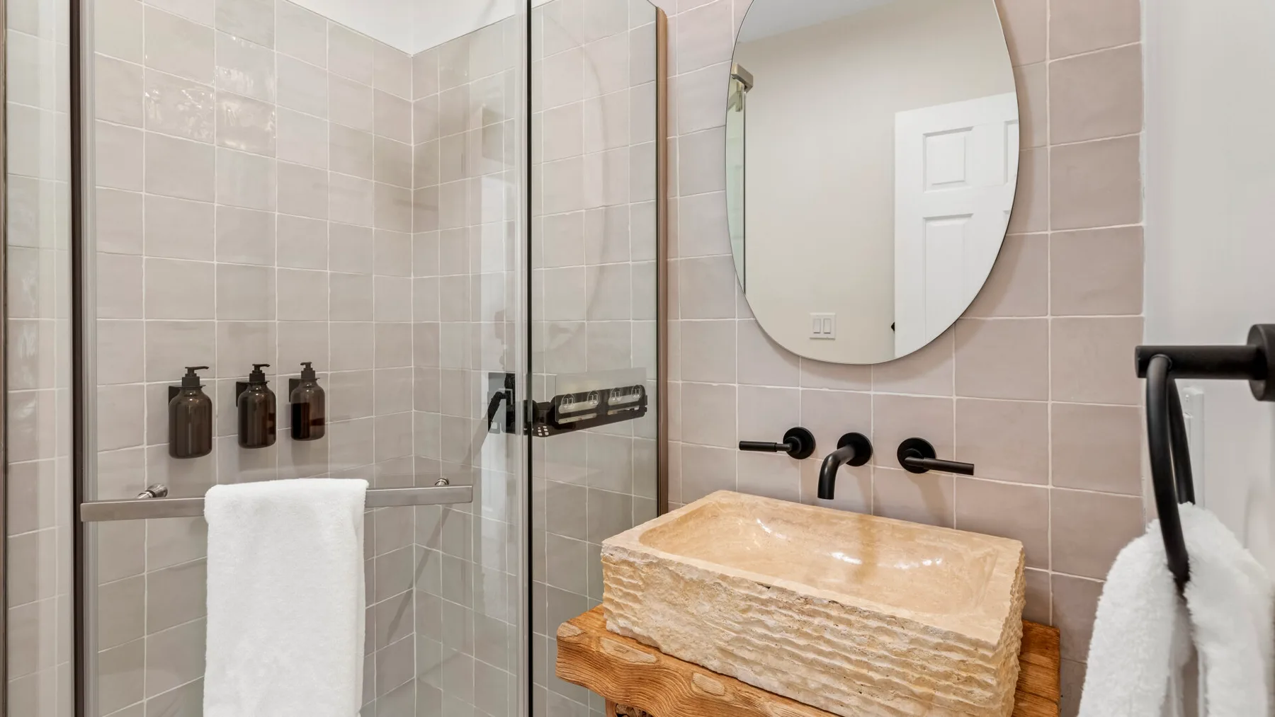 A tiled bathroom features a glass shower and a vessel sink beneath an oval mirror.